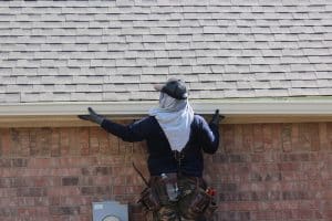 A worker from a gutter company, wearing gloves, a cap, and a tool belt, inspects or repairs the gutter of a brick house, standing with arms raised toward the roofing.
