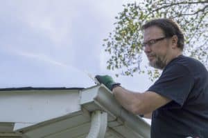 A man wearing glasses and gloves cleans debris from the seamless gutters of a house on a clear day, with green tree branches visible in the background.